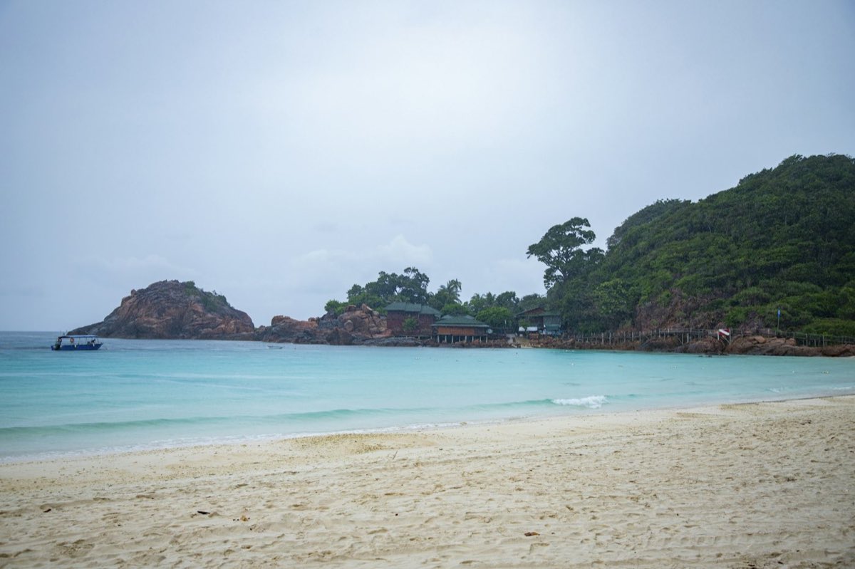 A white-sand beach on Redang Island with a wooded headland and a moored boat
